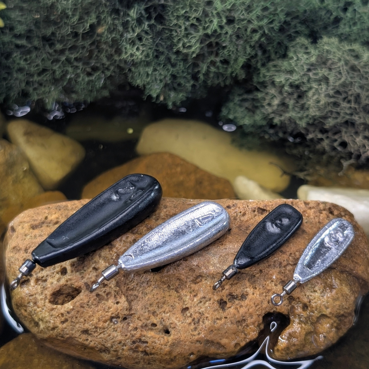 Black and unpainted Free Rig weights with closed swivel eyes displayed on a rock in shallow water.