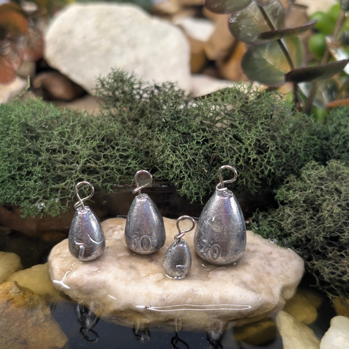 Four unpainted Fastach Jika Rig weights with quick-change attachment loops displayed on a stone in shallow water.