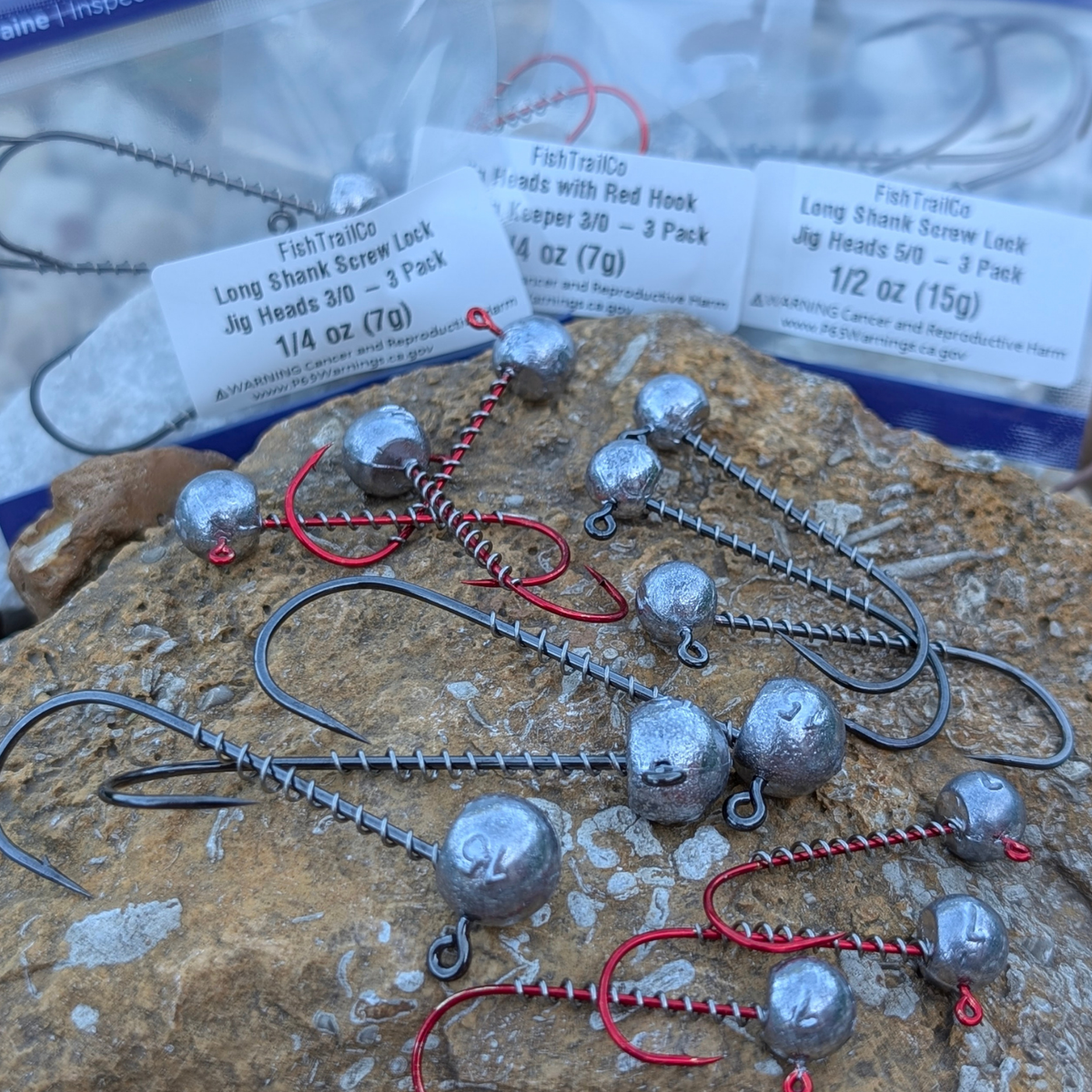 FishTrailCo Long Shank and red hook spring keeper jig heads displayed on a rock in front of their packages.