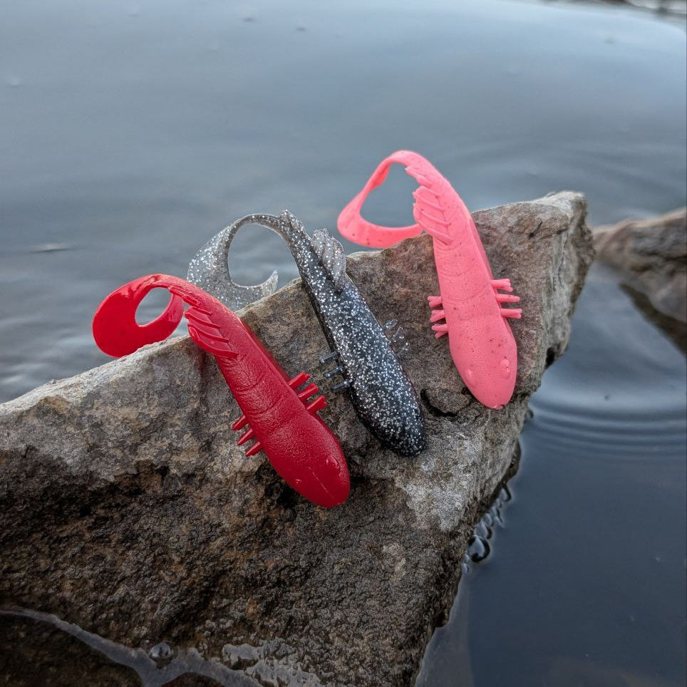 Red, gray, and pink Bait Breath BeTanCo 3-inch curly tail lures resting on a log beside the water.