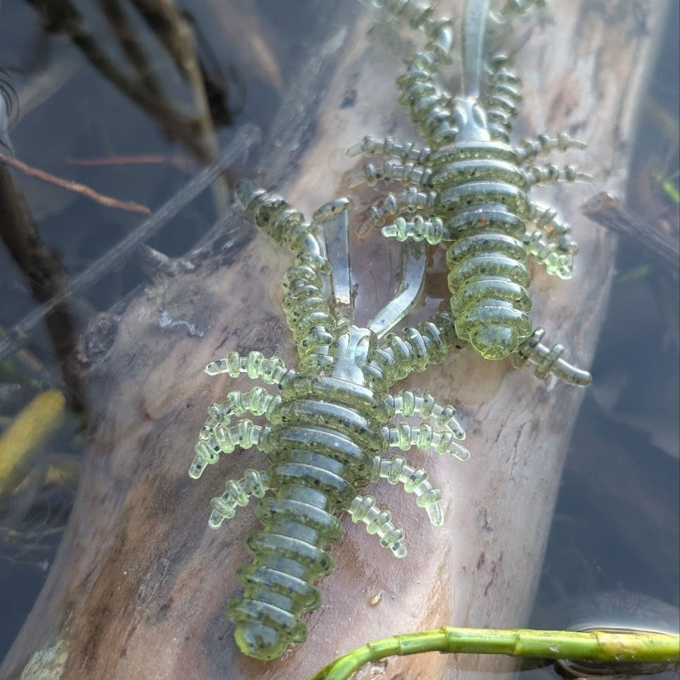 Two olive-green Bait Breath U30 Mosya 3-inch creature baits resting on a log beside the water.